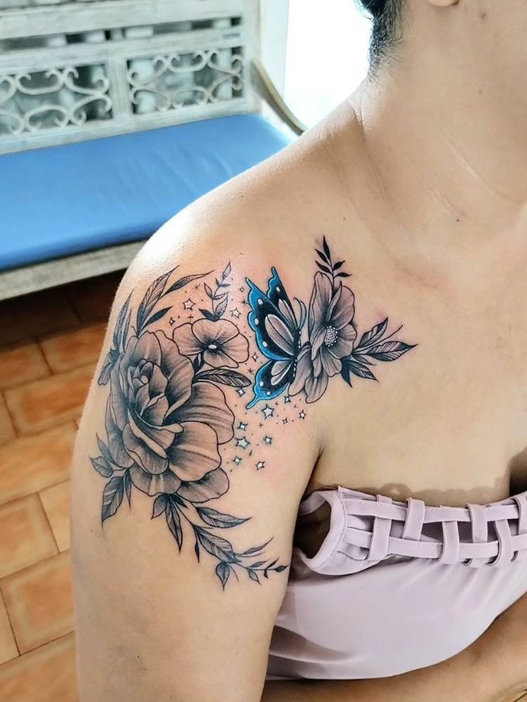 tatuagem-feminina-ombro-flor-borboleta-azul-sombras Mulher de pele clara com blusa rosa antigo e tatuagem de flores sombreadas com borboleta azul no ombro