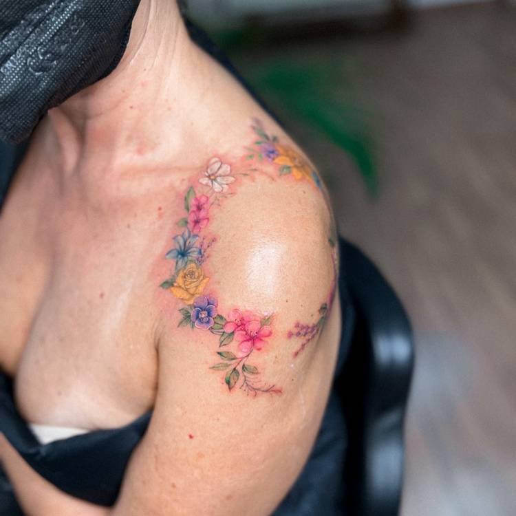 tatuagem-feminina-ombro-flores-coloridas Mulher de pele clara com blusa azul e tatuagem de ramos de flores coloridas ao redor do ombro