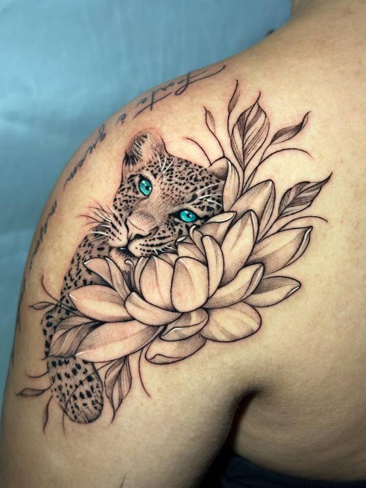 tatuagem-feminina-ombro-onca Mulher de pele clara com onça realista tatuada no ombro, com olhos azuis e abraçada com flores