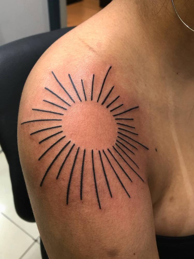 tatuagem-feminina-ombro-raios-solares Mulher de pele morena com tatuagem de sol no ombro, apenas com os raios e círculo vazado, em traço preto grosso