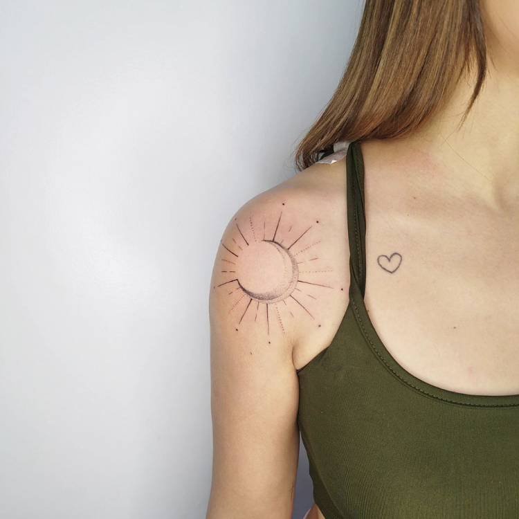 tatuagem-feminina-ombro-sol-2 Mulher de pele clara usando regata verde com tatuagem de coração próxima ao colo e tatuagem de sol fine line no ombro