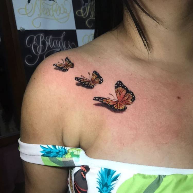 tatuagem-feminina-ombro-sombra-borboleta Mulher de pele clara e blusa verde com tatuagem de três borboletas laranjas de tamanho crescente no ombro