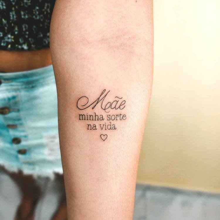 Uma tatuagem no antebraço que diz "Mãe minha sorte na vida" com um pequeno coração abaixo. A palavra "Mãe" está em caligrafia elaborada, enquanto o restante do texto é em fonte simples.