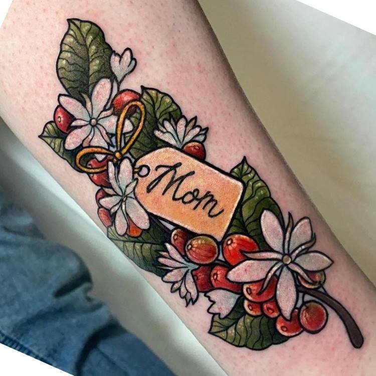 Uma tatuagem vibrante no antebraço, com um ramo de café apresentando folhas verdes escuras, flores brancas e frutos vermelhos. Um tag laranja com "Mom" em caligrafia está amarrado ao ramo, simbolizando carinho e homenagem.