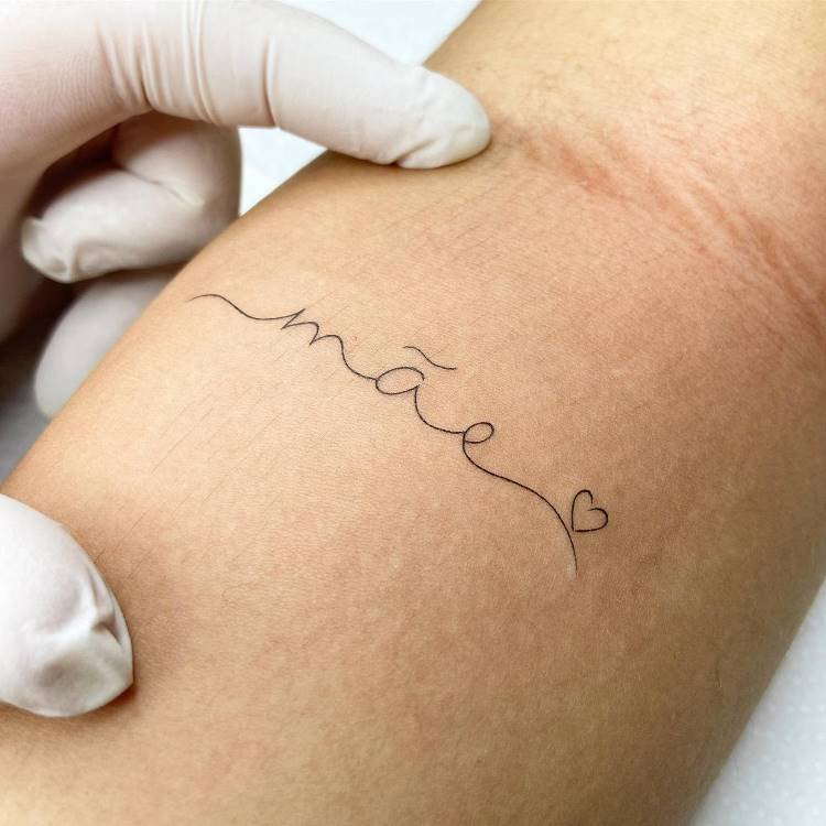 tatuagem-mae-pai-13 A imagem mostra o antebraço de uma pessoa com uma tatuagem fina e delicada da palavra "mãe" em escrita cursiva, seguida por um pequeno coração, ambos em preto. A pele apresenta algumas marcas e uma mão enluvada de branco toca a pele.