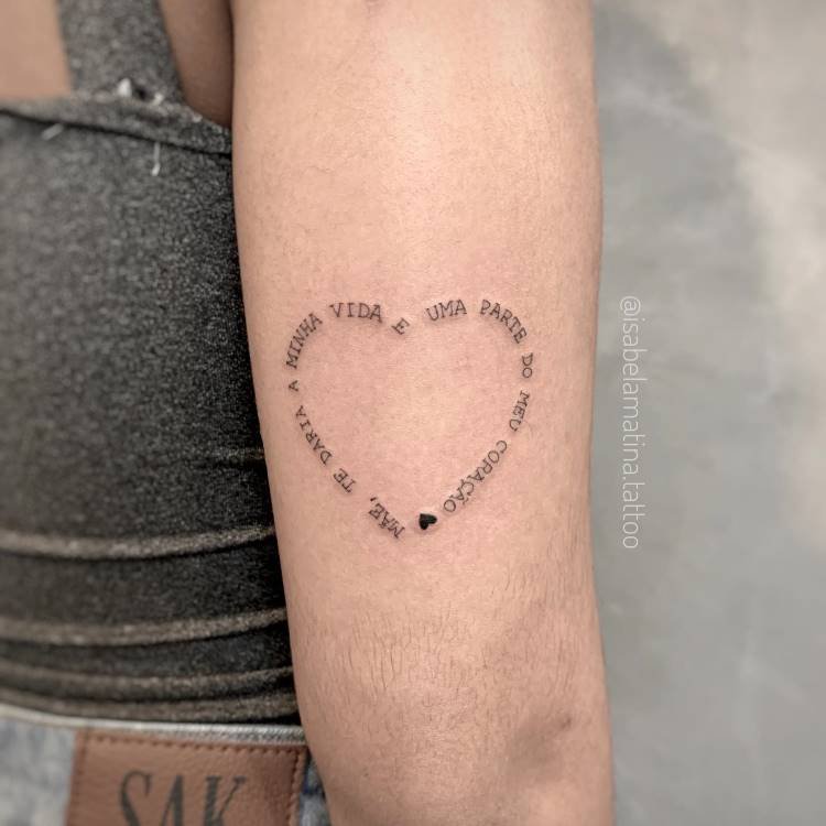 Uma tatuagem de linha fina em formato de coração no antebraço de uma pessoa. As palavras "MÃE, TE DARIA A MINHA VIDA, É UMA PARTE DO MEU CORAÇÃO" estão escritas ao redor do contorno do coração, com um pequeno coração preenchido no final.