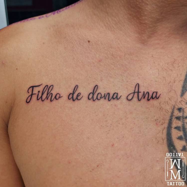 tatuagem-mae-pai-15 A imagem mostra o torso de um homem com uma tatuagem preta no peito que diz "Filho de dona Ana" em caligrafia cursiva. Parte de outra tatuagem preta pode ser vista no ombro direito do homem. No canto inferior direito, há um logotipo em branco com a palavra "TATTOO" invertida.