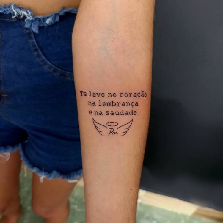 tatuagem-mae-pai-16 A imagem mostra uma tatuagem no antebraço de uma pessoa com a frase "Te levo no coração na lembrança e na saudade.", seguida pela palavra "Pai" e um par de asas de anjo com uma auréola. A tatuagem é em preto e a pele parece clara. A pessoa veste shorts jeans.