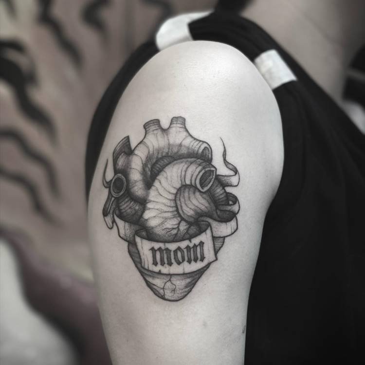 Uma tatuagem de um coração anatômico em preto e branco está no braço. O coração é detalhado com sombras e linhas finas, e uma fita com a palavra "MOM" em fonte gótica o envolve, adicionando um toque gótico ao design.