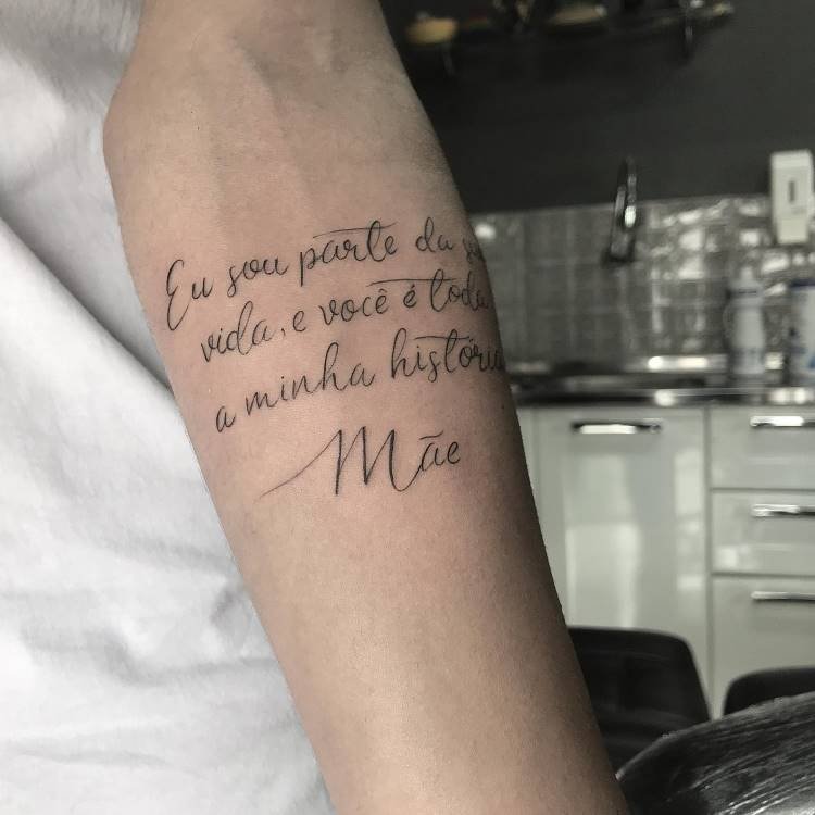 Uma tatuagem no antebraço que diz: "Eu sou parte da vida, e você é toda a minha história. Mãe". A tatuagem é escrita em caligrafia fina e preta, destacando-se na pele clara.