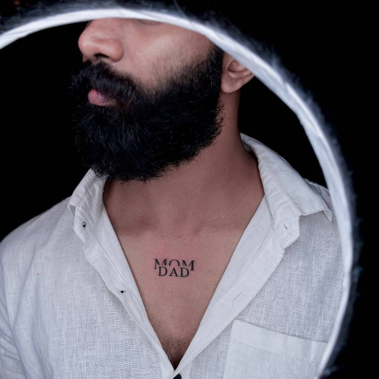 tatuagem-mae-pai-5 A imagem apresenta um close-up de um homem barbudo vestindo uma camisa branca aberta, revelando uma tatuagem preta no centro do peito, que diz "MOM" em cima de "DAD". A iluminação forma um círculo ao redor da cabeça do homem, com o fundo escuro.