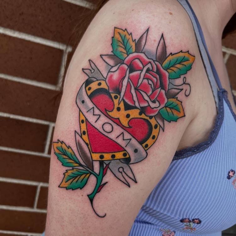 tatuagem-mae-pai-6 A imagem mostra uma tatuagem colorida no braço de uma pessoa, no estilo tradicional. Ela apresenta um coração vermelho e amarelo, perfurado por um punhal e com uma faixa cinza escrito "MOM". Uma rosa vermelha e folhas verdes e amarelas adornam o desenho.