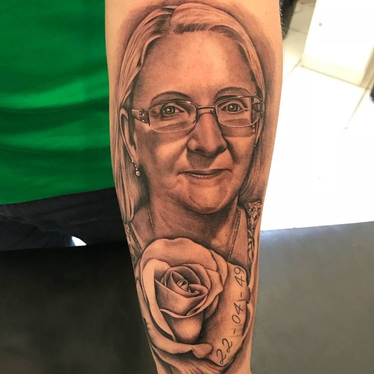 Uma tatuagem realista em preto e branco de uma mulher com óculos no antebraço. Abaixo do retrato, uma rosa grande com a data "22-04-49" gravada em uma de suas pétalas.