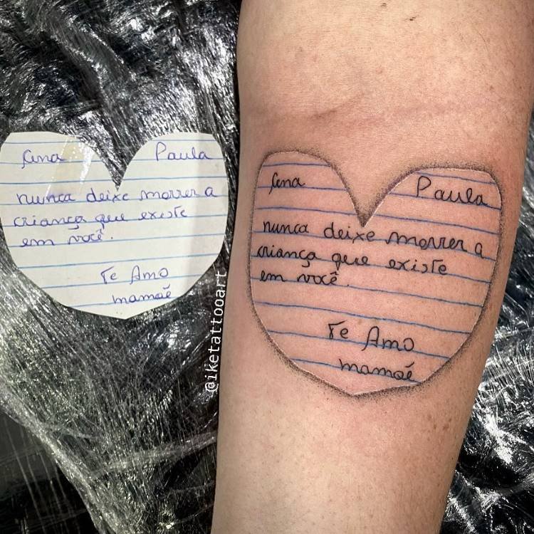 Uma tatuagem em formato de coração imita um bilhete de papel pautado no antebraço esquerdo de uma pessoa. O texto manuscrito diz: "lina Paula nunca deixe morrer a criança que existe em você. Te Amo mamãe". Uma cópia do bilhete original está ao lado, realçando a semelhança.