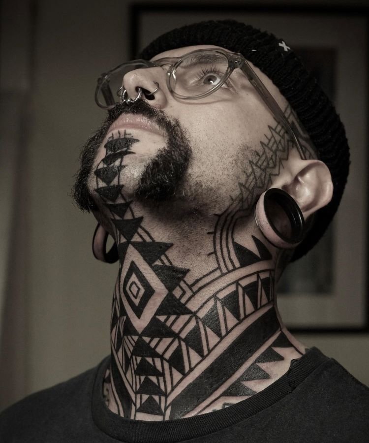tatuagem-masculina-no-pescoco-10 A imagem mostra uma pessoa com óculos, barba e alargadores nas orelhas. Ela tem uma tatuagem no pescoço de padrões geométricos em preto, que cobrem desde o queixo até a clavícula. As formas incluem triângulos, losangos e linhas, criando um design tribal intrincado e simétrico.
