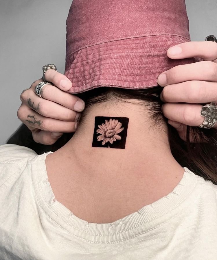 tatuagem-masculina-no-pescoco-19 A imagem mostra uma pessoa de costas usando um chapéu rosa. Ela tem uma tatuagem no pescoço de uma flor detalhada dentro de um quadrado preto. A pessoa está segurando o chapéu com ambas as mãos, revelando vários anéis nos dedos. A tatuagem é centralizada na nuca.