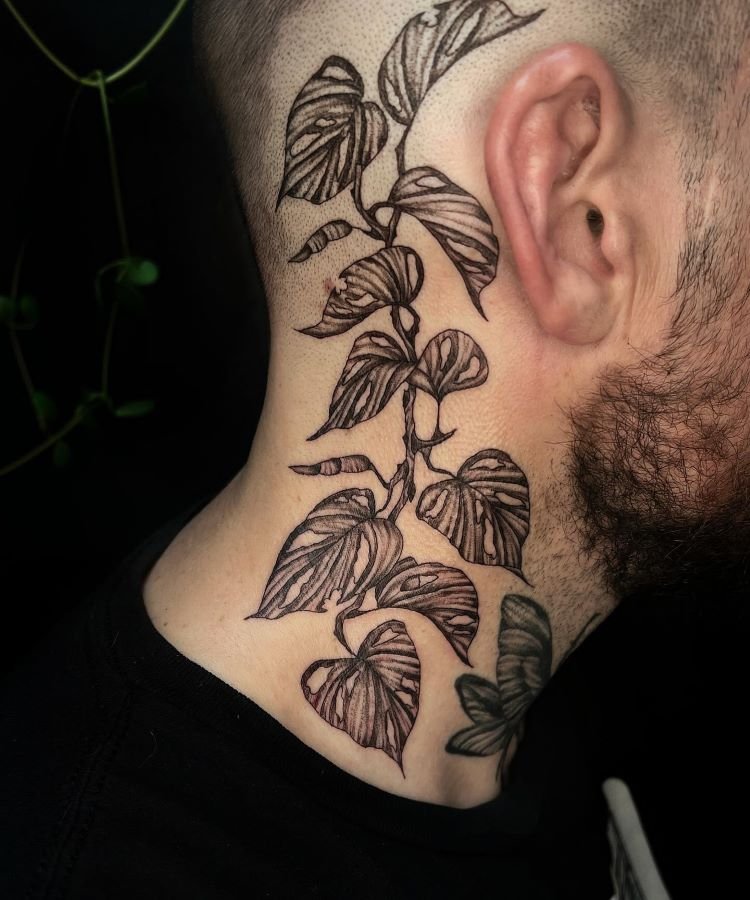 tatuagem-masculina-no-pescoco-21 A imagem mostra uma pessoa de perfil com uma tatuagem no pescoço de uma planta trepadeira com folhas detalhadas em preto. A pessoa tem barba e usa uma camiseta preta. A tatuagem se estende da base do pescoço até atrás da orelha.