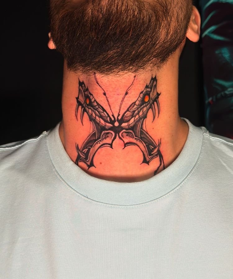 tatuagem-masculina-no-pescoco-24 A imagem mostra uma pessoa com barba e camiseta clara. Ela tem uma tatuagem no pescoço de um design simétrico e abstrato em preto, com detalhes em vermelho. A tatuagem parece ter duas cabeças de dragão opostas, com garras e olhos brilhantes, cobrindo a parte frontal do pescoço.