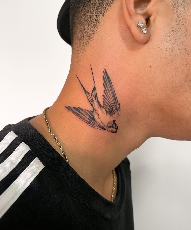 tatuagem-masculina-no-pescoco-3 A imagem mostra uma pessoa de perfil com cabelo curto e uma corrente dourada. Ela tem uma tatuagem no pescoço de uma andorinha em voo, desenhada em preto e cinza, com detalhes realistas nas asas e no corpo. A pessoa usa brincos brilhantes e uma camiseta preta com listras brancas.