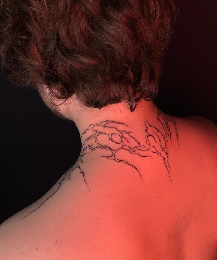 tatuagem-masculina-no-pescoco-6 A imagem mostra uma pessoa de costas com cabelo castanho ondulado. Ela tem uma tatuagem no pescoço de galhos finos e detalhados, desenhados em linhas pretas que se estendem da base do crânio até os ombros. A tatuagem é delicada e parece parte de uma árvore.