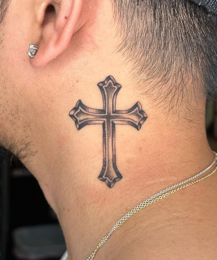 tatuagem-masculina-no-pescoco-7 A imagem mostra uma pessoa de perfil usando um brinco pequeno e brilhante. Ela tem uma tatuagem no pescoço de uma cruz detalhada em preto, localizada logo abaixo da orelha. A pessoa também usa um colar fino de corrente dourada.