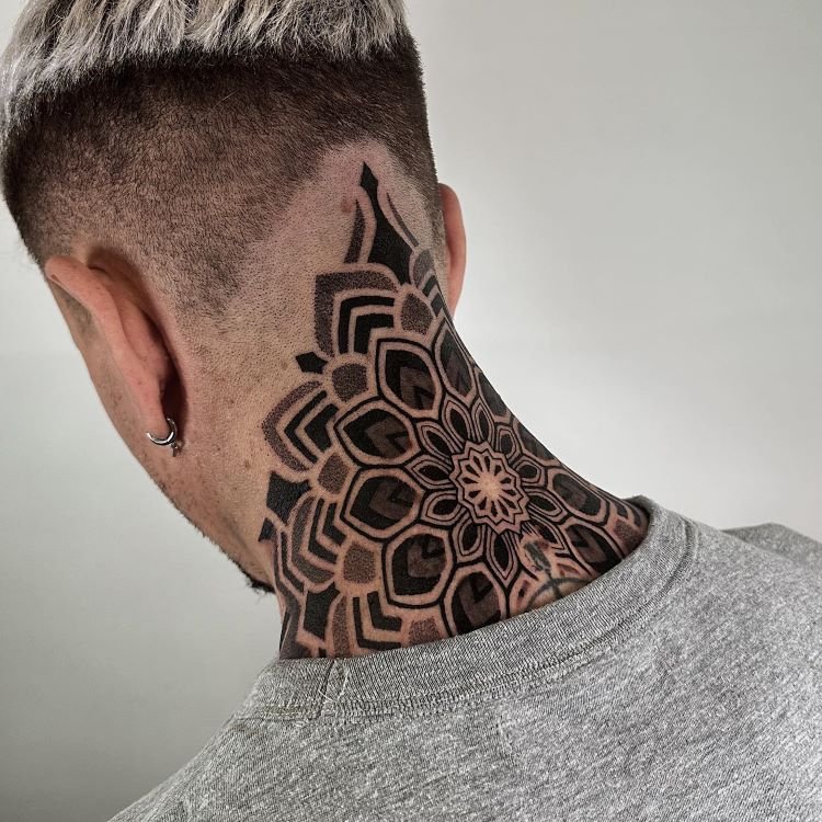 tatuagem-masculina-pescoco-2-1 A imagem mostra uma pessoa de costas com cabelo curto raspado nas laterais. Ela tem uma tatuagem no pescoço de um mandala geométrico detalhado em preto, que se estende da base do crânio até a parte superior das costas. A tatuagem é simétrica e intricada.