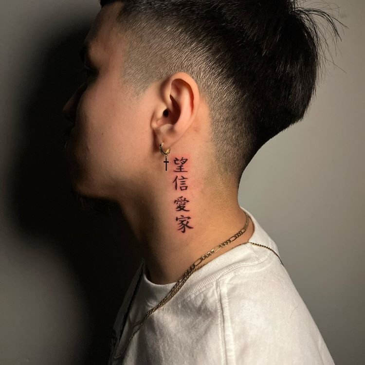 tatuagem-masculina-pescoco-6-1 A imagem mostra uma pessoa de perfil com cabelo curto. Ela tem uma tatuagem no pescoço de caracteres chineses verticais em preto, significando "esperança", "fé", "amor" e "família". A tatuagem está localizada na lateral do pescoço, perto da orelha, onde a pessoa usa um brinco de cruz.