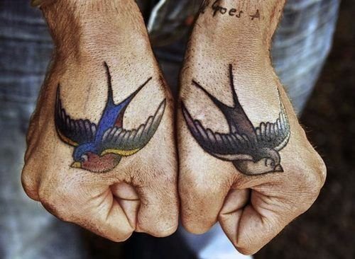 Homem de pele clara com tatuagem de andorinha old school colorida em uma mão e preta na outra