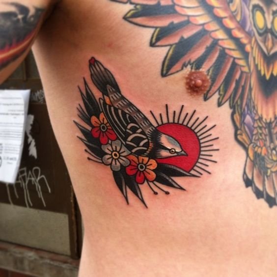 Pessoa de pele clara com tatuagens old school no peitoral e passarinho com flores e sol old school na costela