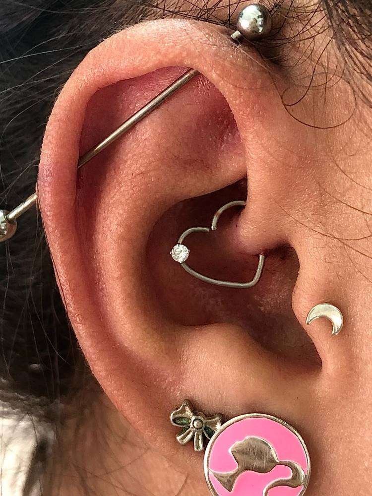 Orelha com piercing transversal, argola de coração no daith, lua no tragus e brinco da Barbie no lóbulo.